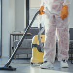 A cleaner in protective coveralls and gloves vacuuming a modern living room.